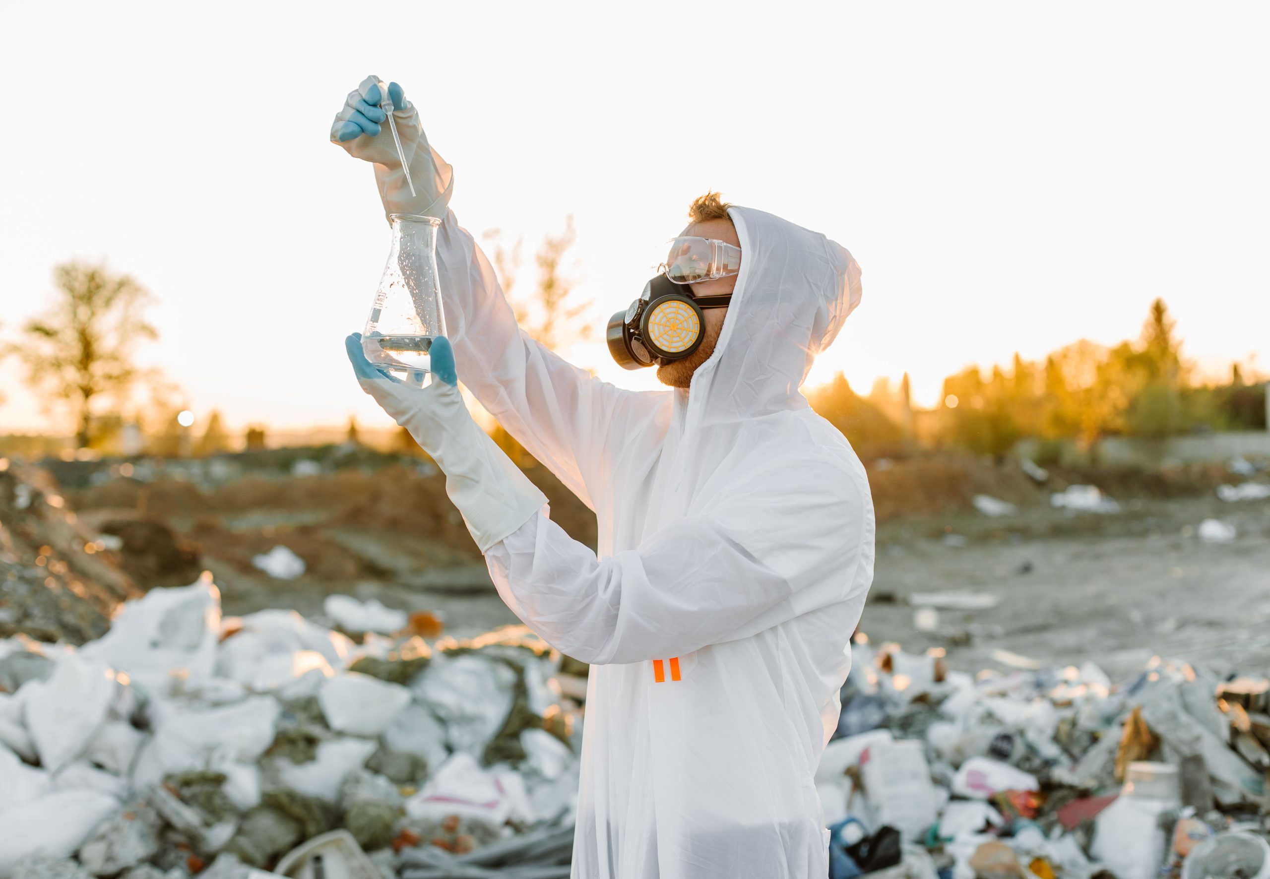 Scientist in protective gear collecting samples at a waste site during sunset.